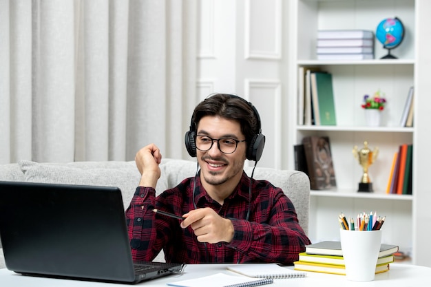 Estudiante en línea chico lindo en camisa a cuadros con gafas estudiando en la computadora sonriendo y sosteniendo la pluma