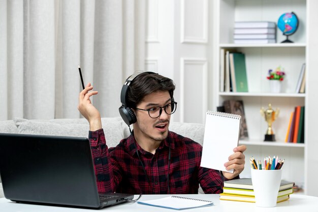 Estudiante en línea chico lindo en camisa a cuadros con gafas estudiando en la computadora sorprendido por las notas