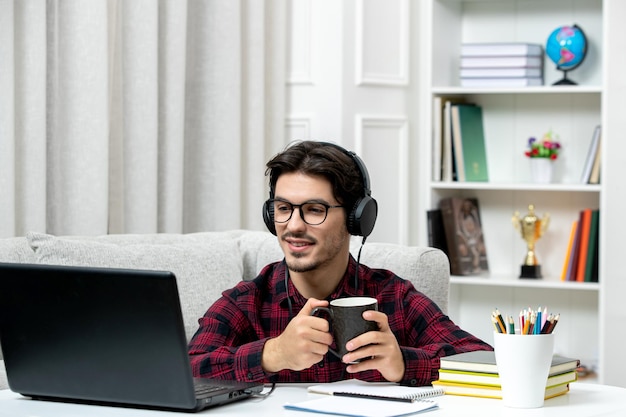 Estudiante en línea chico lindo en camisa a cuadros con gafas estudiando en la computadora con taza de café