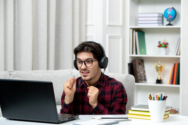 Estudiante en línea joven en camisa a cuadros con gafas estudiando en la computadora levantando los puños