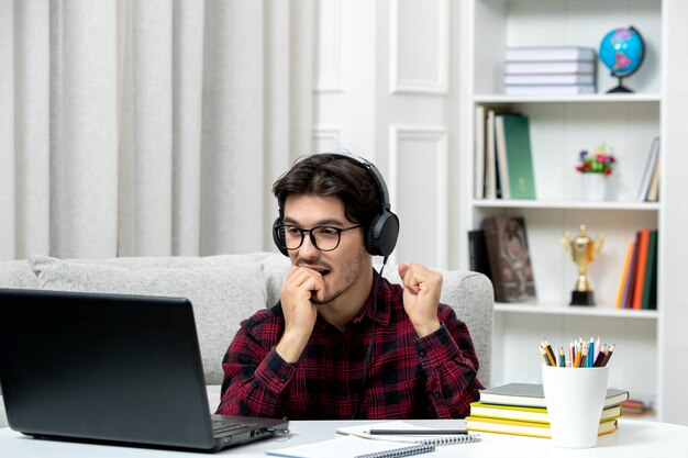 Estudiante en línea joven en camisa a cuadros con gafas estudiando en la computadora mordiendo el dedo