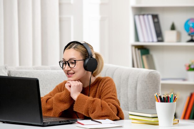 Estudiante en línea joven linda chica con gafas y suéter naranja estudiando en la computadora sonriendo felizmente