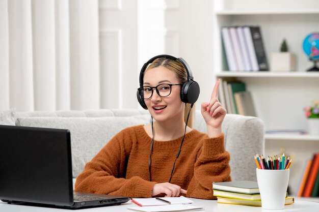 Estudiante en línea joven linda chica con gafas y suéter naranja estudiando en la computadora sonriendo