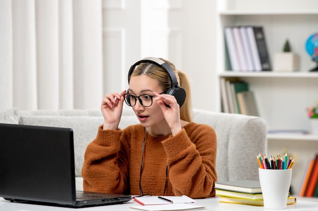 Foto gratuita estudiante en línea joven linda chica en gafas y suéter naranja estudiando en computadora sorprendido