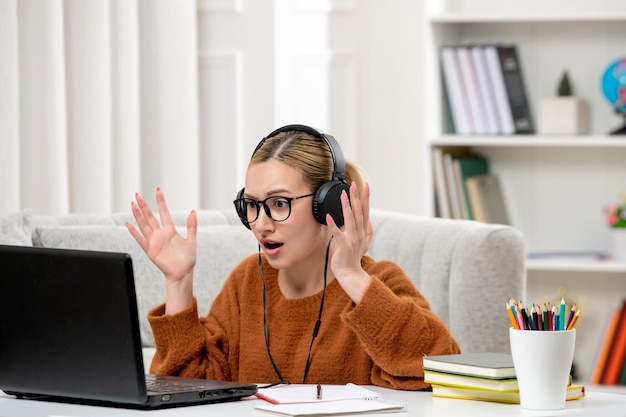 Estudiante en línea linda chica con gafas y suéter estudiando en la computadora con auriculares confundidos