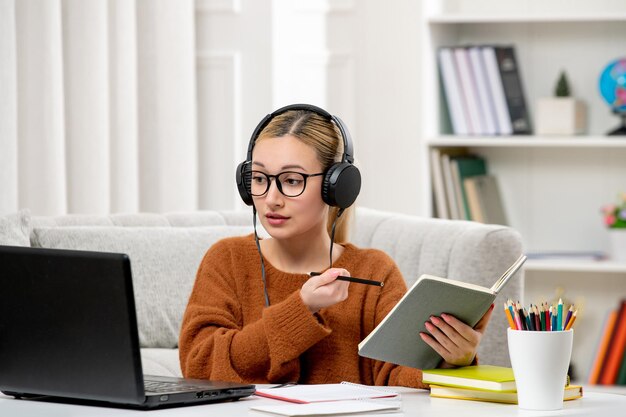 Estudiante en línea linda chica con gafas y suéter estudiando en la computadora escribiendo notas