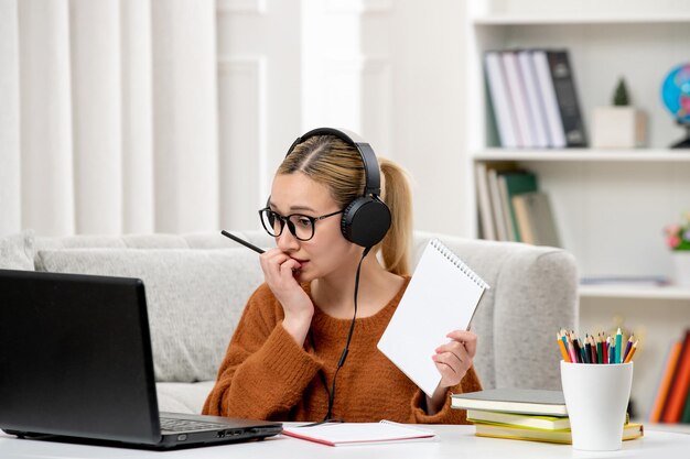 Estudiante en línea linda chica con gafas y suéter estudiando en la computadora mordiendo el dedo