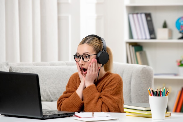 Estudiante en línea linda chica con gafas y suéter estudiando en la computadora sorprendida en los auriculares