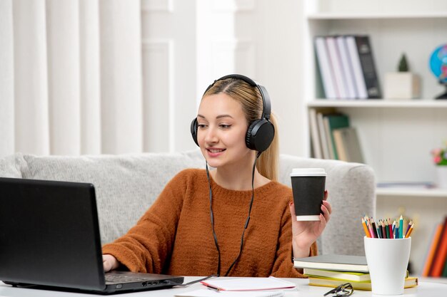 Estudiante en línea linda chica con gafas y suéter estudiando en la computadora sosteniendo un vaso de papel