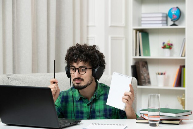 Estudiante en línea lindo joven estudiando en la computadora con gafas en camisa verde con bloc de notas de pluma