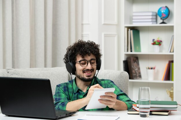 Estudiante en línea lindo joven estudiando en la computadora con gafas en camisa verde feliz tomando notas