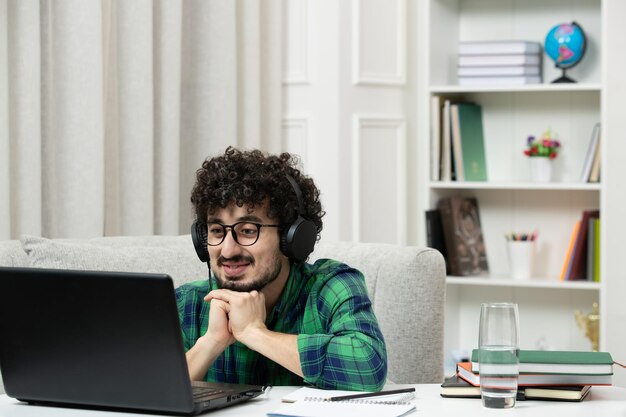 Estudiante en línea lindo joven estudiando en la computadora con gafas en camisa verde sonriendo y esperando