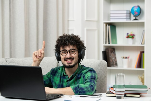 Estudiante en línea lindo joven estudiando en la computadora con gafas en camisa verde sonriendo y señalando