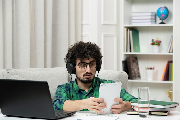 Estudiante en línea lindo joven estudiando en computadora con gafas camisa verde sorprendido leyendo notas