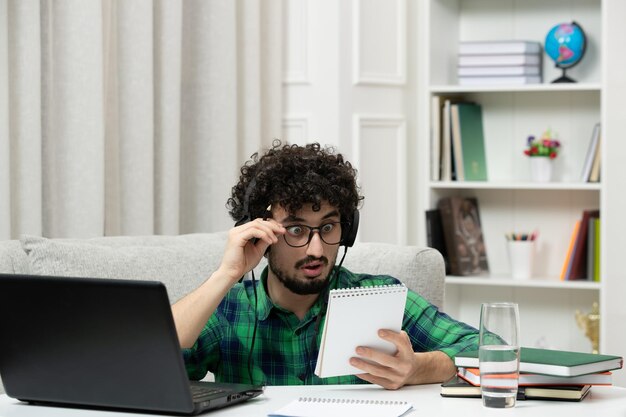 Estudiante en línea lindo joven estudiando en computadora con gafas en camisa verde sorprendido leyendo