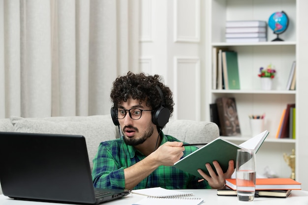 Estudiante en línea lindo joven estudiando en la computadora con gafas en camisa verde sosteniendo un bloc de notas