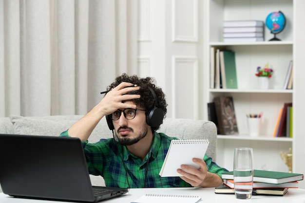 Estudiante en línea lindo joven estudiando en computadora con gafas en camisa verde sosteniendo la cabeza