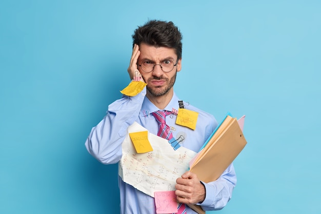 El estudiante masculino sin afeitar grave e intenso tiene dolor de cabeza debido a un largo trabajo ocupado preparándose para el examen de matemáticas o hace que el proyecto tenga fecha límite lleva trabajos con sumas escritas