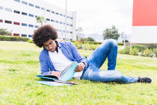 Estudiante masculino africano africano joven que pone en la hierba verde que lee el libro