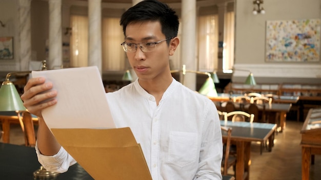 Estudiante masculino asiático abriendo un sobre con los resultados de los exámenes en la biblioteca universitaria