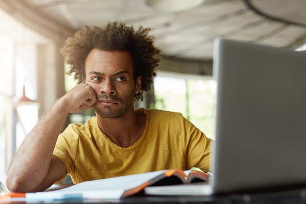 Estudiante masculino con cabello tupido y piel oscura con expresión aburrida de estar agotado de estudiar sentado en un café rodeado de libros y un cuaderno haciendo su proyecto de investigación usando internet gratis