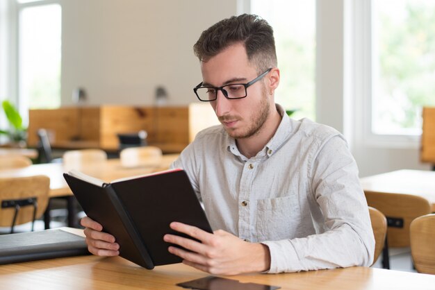 Estudiante masculino elegante que lee el libro de texto en el escritorio en sala de clase