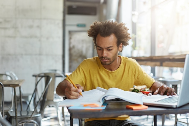 Estudiante masculino europeo negro concentrado con barba preparándose para la prueba de examen, sentado en el comedor de la universidad, comiendo sándwich, buscando información en Internet, usando una computadora portátil