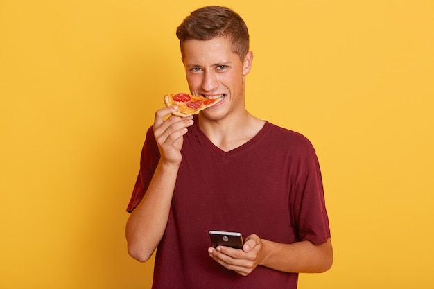 Estudiante masculino guapo mordiendo deliciosa rebanada de pizza, vistiendo camiseta burdeos, de pie contra la pared amarilla