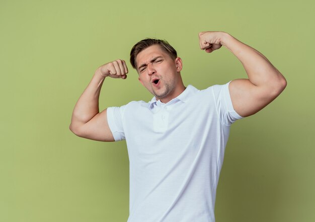 Estudiante masculino hermoso joven alegre que hace gesto fuerte aislado en fondo verde oliva