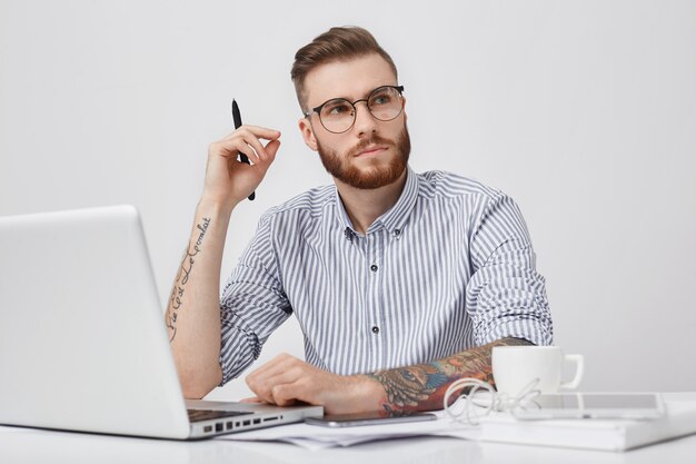 Estudiante masculino inteligente pensativo con haido de moda mira pensativamente a un lado mientras intenta reunir pensamientos, trabaja en el papel del curso, se sienta frente a la computadora portátil abierta