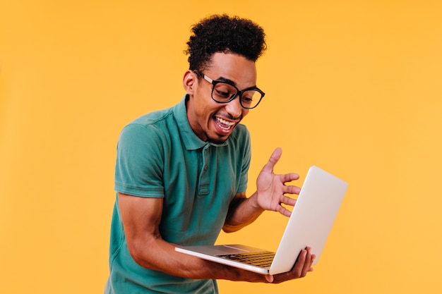 Estudiante masculino rizado sorprendido que mira la pantalla del ordenador portátil. Filmación en interiores de freelancer africano lleva gafas.