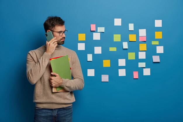 estudiante masculino serio lee publicaciones pegajosas en la pared azul, gira a la derecha tiene conversación telefónica sostiene libros de texto coloridos vestidos casualmente discute la preparación del examen.