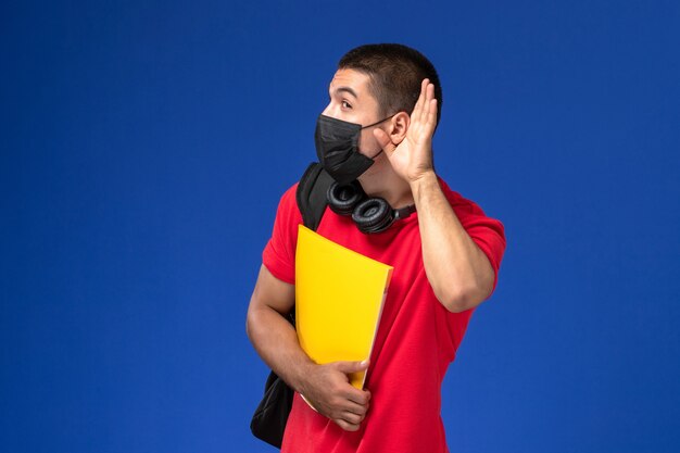Estudiante masculino de vista frontal en camiseta roja con máscara con mochila sosteniendo un archivo amarillo tratando de escuchar sobre fondo azul.