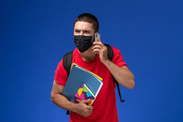 Estudiante masculino de vista frontal en camiseta roja con mochila con máscara con archivos de cuaderno hablando por teléfono en el escritorio azul.