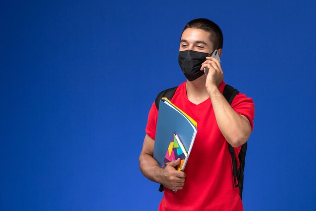 Estudiante masculino de vista frontal en camiseta roja con mochila con máscara con archivos de cuaderno hablando por teléfono sobre el fondo azul.