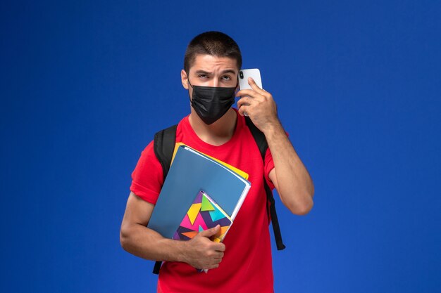 Estudiante masculino de vista frontal en camiseta roja con mochila con máscara con archivos de cuaderno hablando por teléfono sobre el fondo azul.