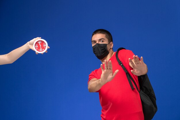 Estudiante masculino de vista frontal en camiseta roja con mochila con máscara asustada de los relojes en el escritorio azul.