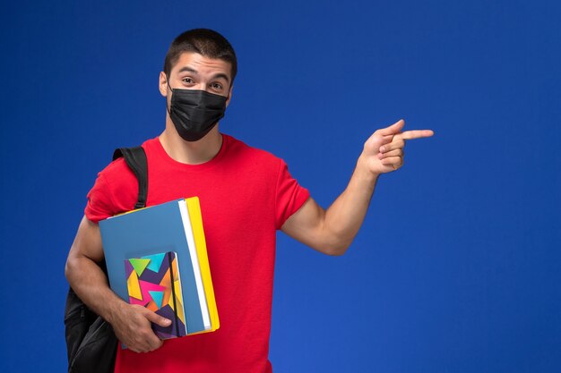 Estudiante masculino de vista frontal en camiseta roja con mochila en máscara estéril negra con archivos sobre el fondo azul.