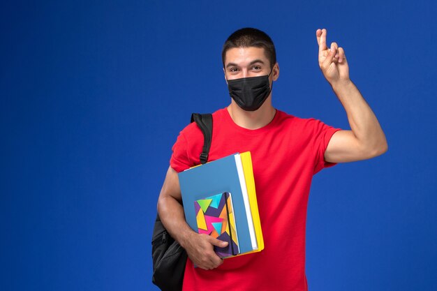 Estudiante masculino de vista frontal en camiseta roja con mochila en máscara estéril negra con cuaderno y archivos sobre el fondo azul.