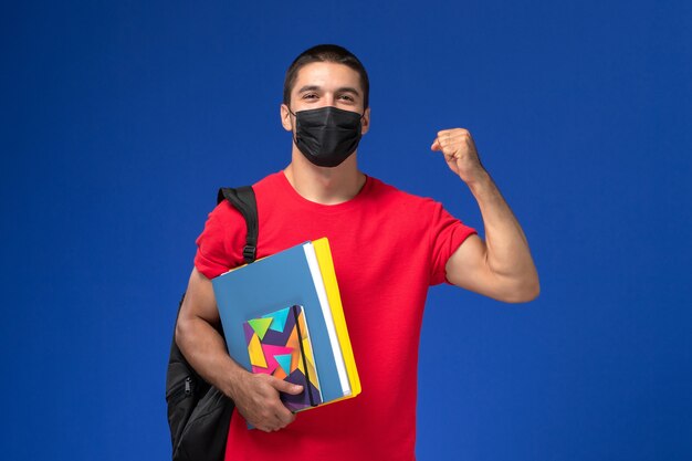 Estudiante masculino de vista frontal en camiseta roja con mochila en máscara estéril negra con cuaderno y archivos sobre el fondo azul.