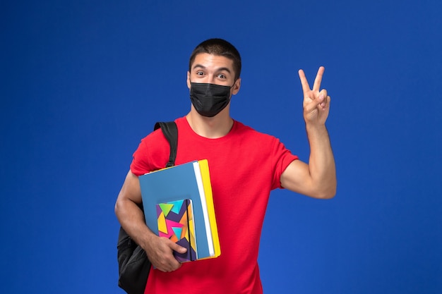 Estudiante masculino de vista frontal en camiseta roja con mochila en máscara estéril negra con cuaderno y archivos sobre el fondo azul.