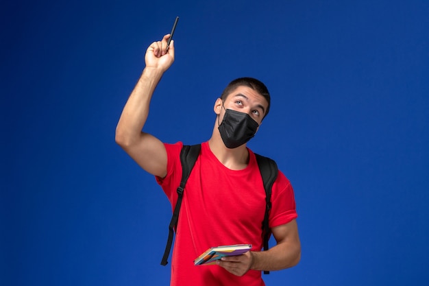 Estudiante masculino de vista frontal en camiseta roja con mochila en máscara estéril negra con lápiz y cuaderno sobre fondo azul.