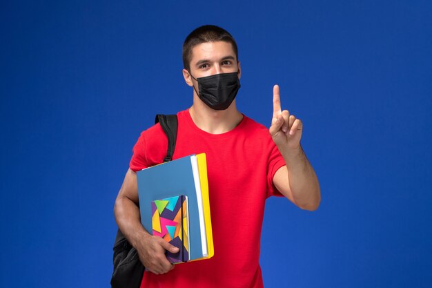 Estudiante masculino de vista frontal en camiseta roja con mochila en máscara estéril negra sosteniendo archivos con el dedo levantado sobre el fondo azul.