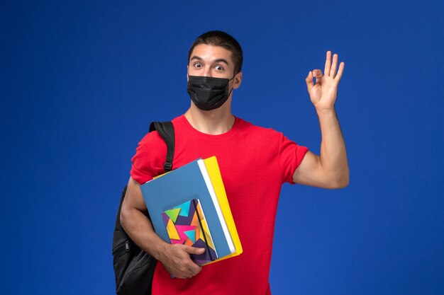 Estudiante masculino de vista frontal en camiseta roja con mochila en máscara estéril negra sosteniendo cuadernos sobre el fondo azul.