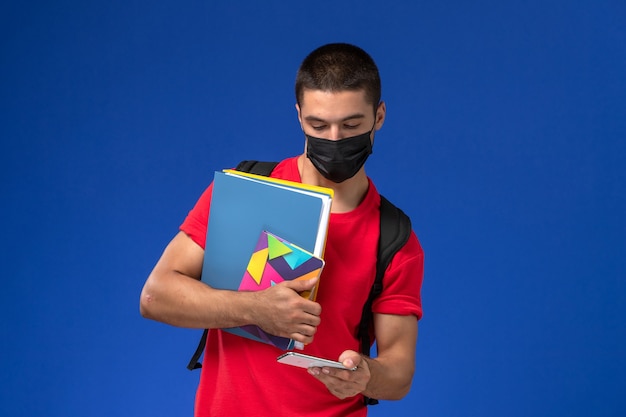 Estudiante masculino de vista frontal en camiseta roja con mochila con máscara sosteniendo archivos y usando su teléfono en el fondo azul.