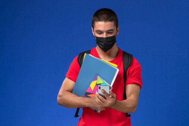 Estudiante masculino de vista frontal en camiseta roja con mochila con máscara sosteniendo archivos y usando el teléfono sobre fondo azul.