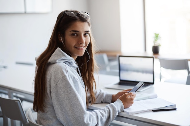 Estudiante de medicina que tiene un día libre tratando de adquirir nuevas habilidades para invertir su tiempo en una nueva empresa Chica usando una computadora portátil y un teléfono mirando a la cámara sonriendo