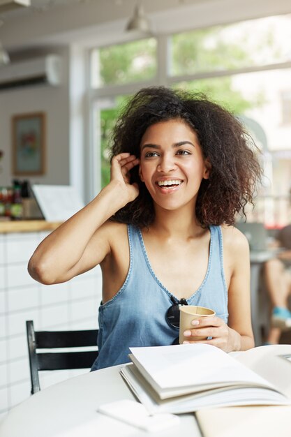 Estudiante de mujer africana alegre hermosa joven que sonríe riendo sentado en café. Libros revistas sobre la mesa. Aprendizaje y educación.