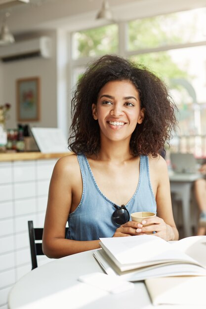 Estudiante de mujer africana alegre hermosa joven que sonríe riendo sentado en café. Libros revistas sobre la mesa. Aprendizaje y educación.