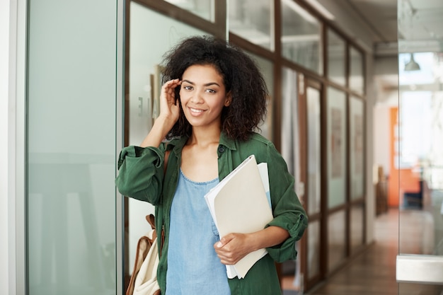 Estudiante de mujer hermoso africano alegre que sonríe el pelo conmovedor que sostiene los libros en universidad. Concepto de educación